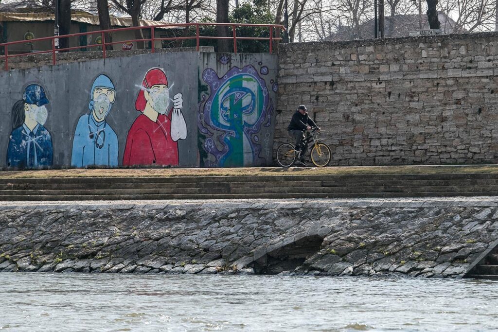 Фотографија од кејот на реката Вардар, на која се гледаат отвори за одлив на водата во реката. Во позадината човек вози велосипед, а на ѕидот има графити.