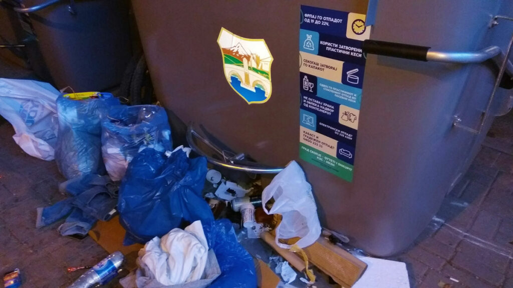 Контејнер со налепница со термини за одложување отпад, со расфрлано ѓубре покрај него.
