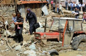 Фотографија на која се гледаат жители кои пешачат преку расфрлани предмети и купишта ѓубре после поплава, се гледа трактор кој е до пола потонат во кал после поплава.